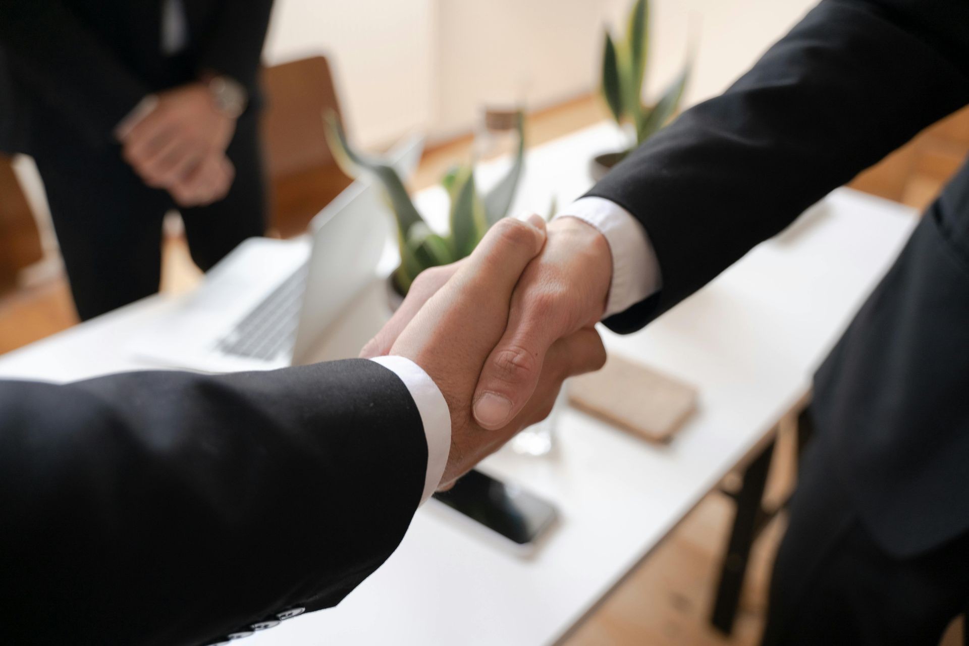 Two people in suits shaking hands over a desk with a laptop and potted plants.